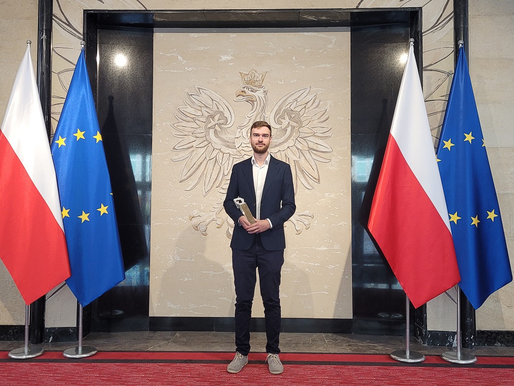 Grzegorz Schnotale ubrany w garnitur pozuje do zdjęcia ze statuetką w rękach, w tle duży orzeł RP, a po prawej i lewej stronie laureata flagi Polski i UE