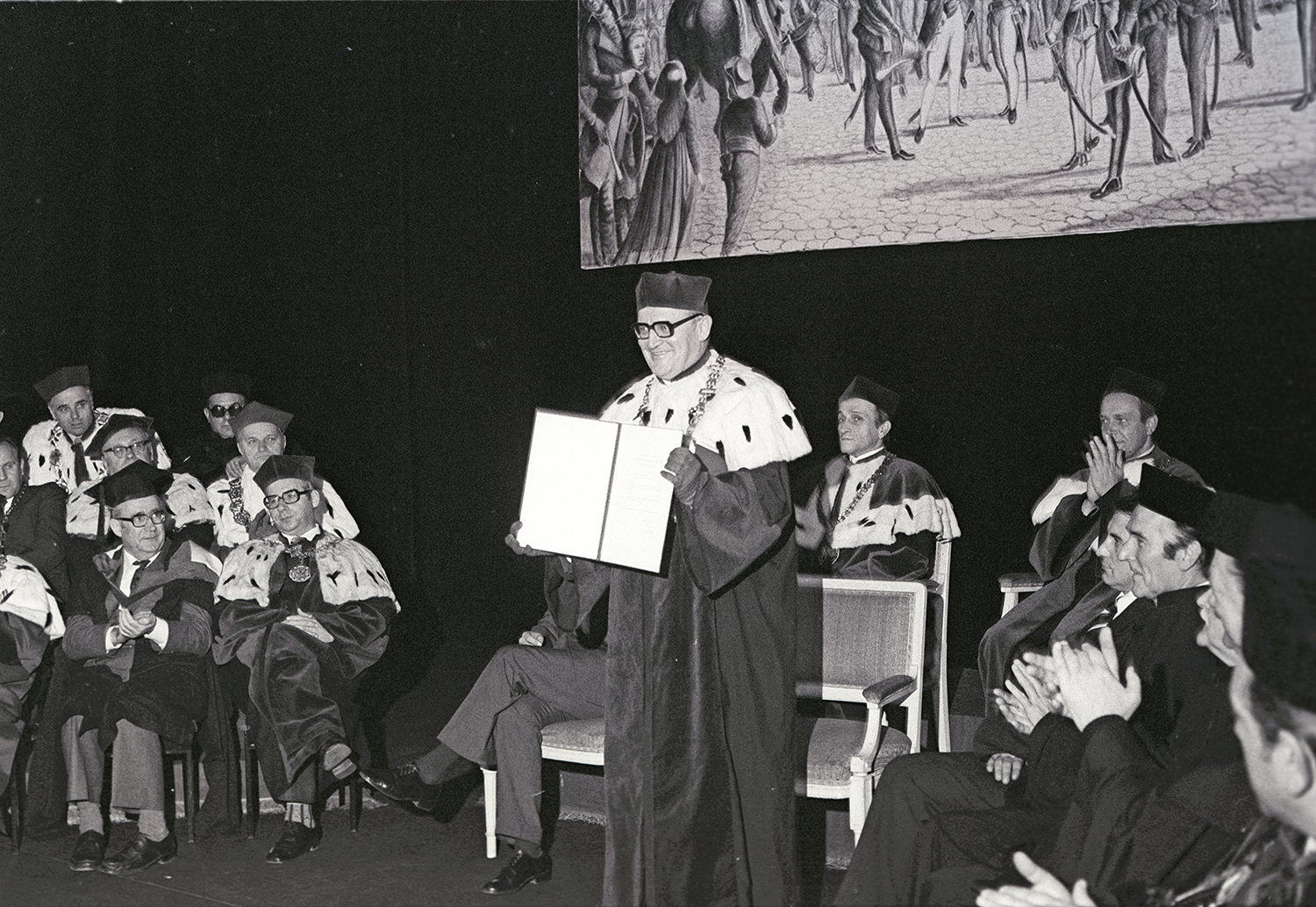 A black-and-white photograph showing a group of university rectors and deans in ceremonial robes. In the center stands a smiling university rector, holding up a document.