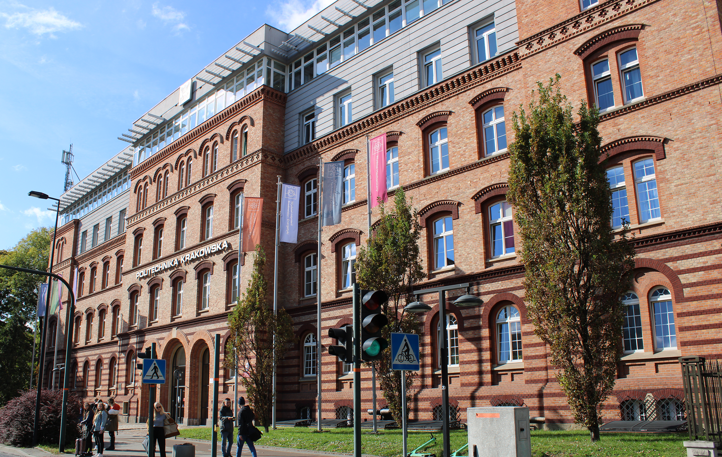 Budynek z brązowej cegły w stylu austro-węgierskim. wzdłuż fasady na wysokich masztach wiszą kolorowe flagi. To budynek główny Politechniki Krakowskiej.