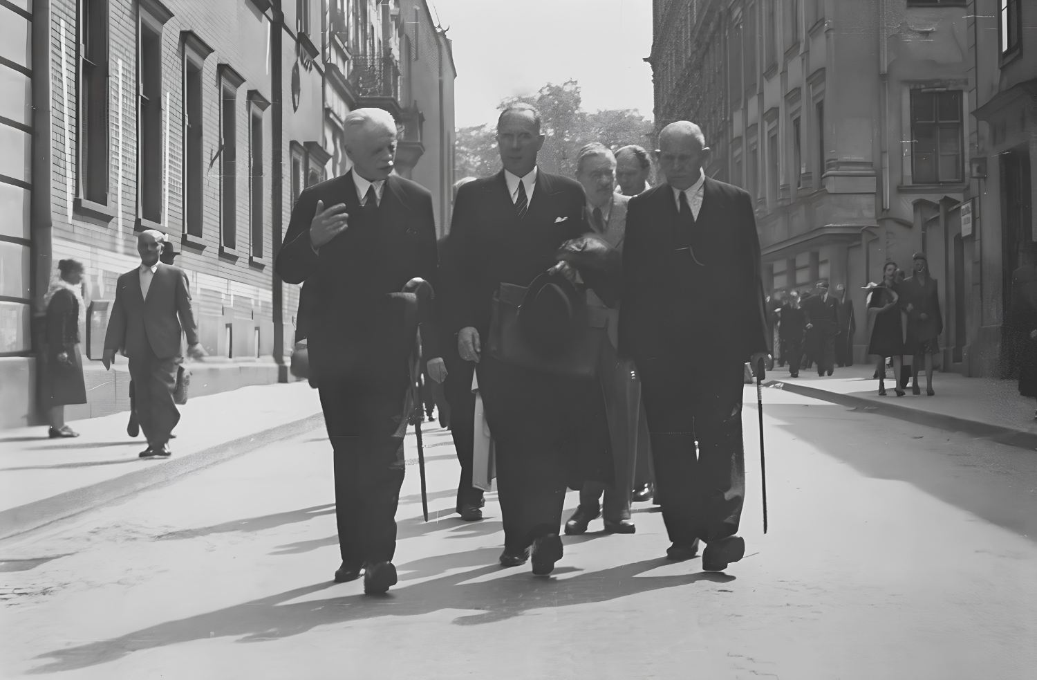 An old black-and-white photograph. A group of people is walking down a city street, led by three elegant men in suits carrying canes. They are Professor Izydor Stella-Sawicki, Rector Walery Goetel, and Professor Jan Krauze.