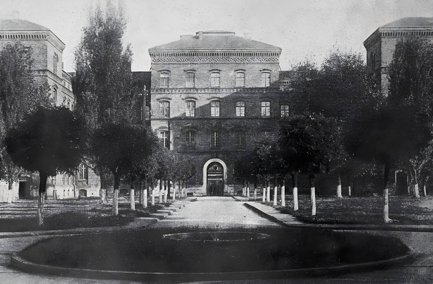 An old black-and-white photograph showing a large brick barracks building and a courtyard with a fountain in the center. Trees grow along the courtyard. This is the headquarters of the Kraków University of Technology in 1945, formerly military barracks.