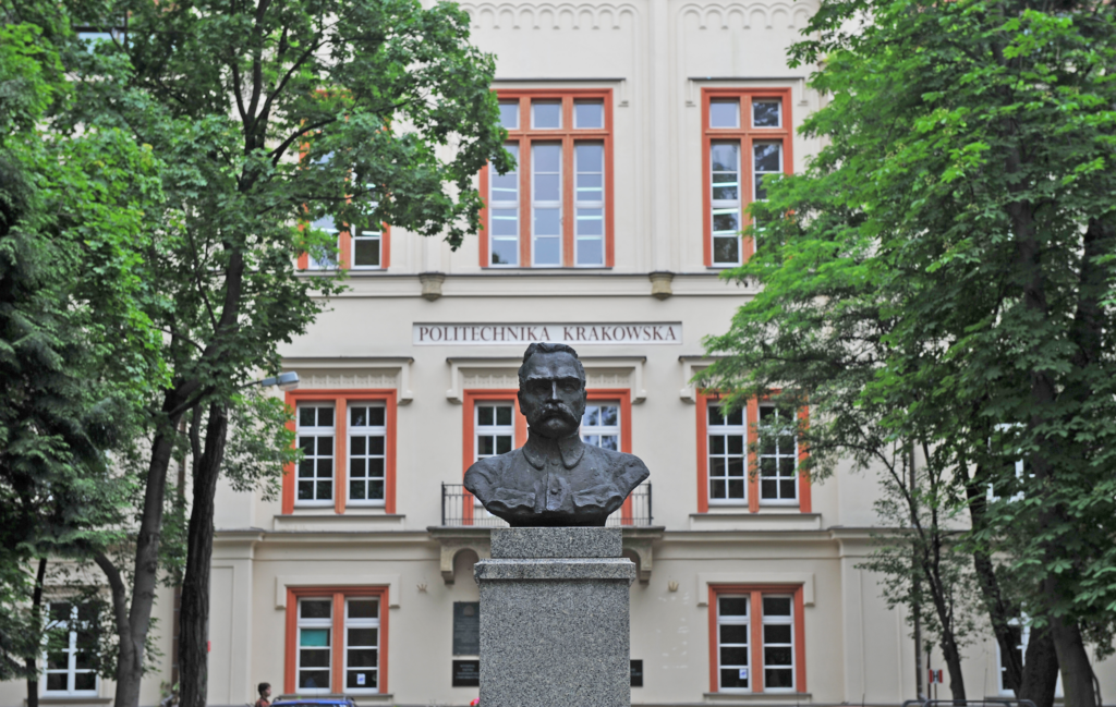 Building of the Kraków University of Technology in Łobzów. Palace façade of light-coloured material with brown window frames. It is surrounded by green trees, and in the foreground a bust of Józef Piłsudski is visible.