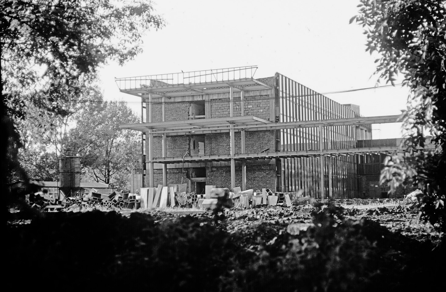 A black-and-white photograph showing a construction site. The photo shows a three-story brick building that does not yet have windows. One wall of the building is entirely glass.
