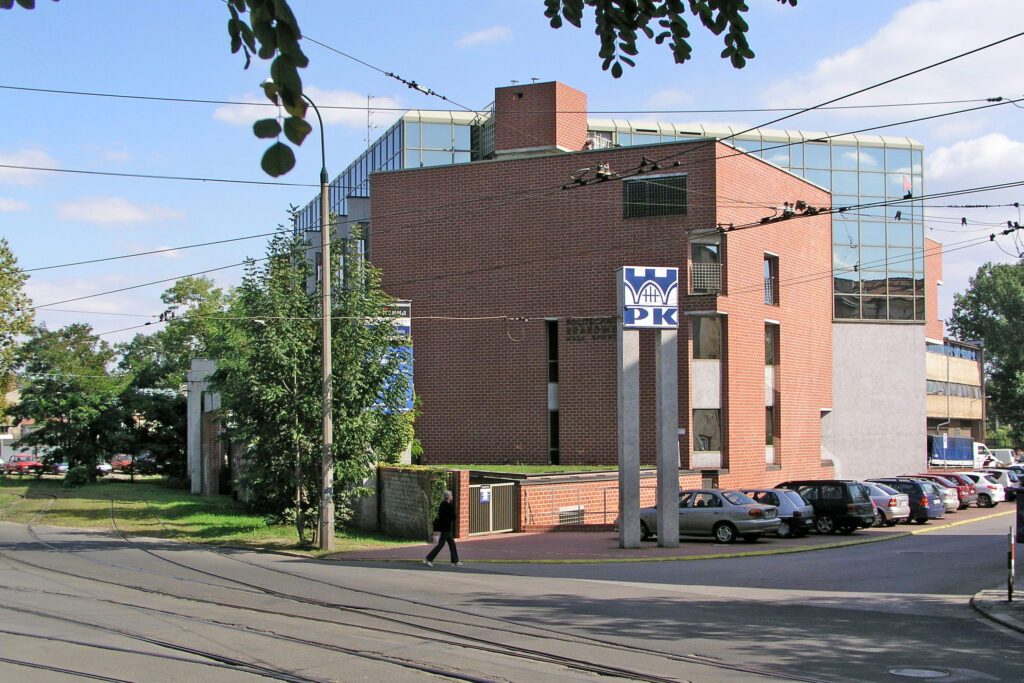 Budynek o nieregularnym kształcie z czerwonej cegły. Przed nim na betonowych filarach stoi sygnet Politechniki Krakowskiej.