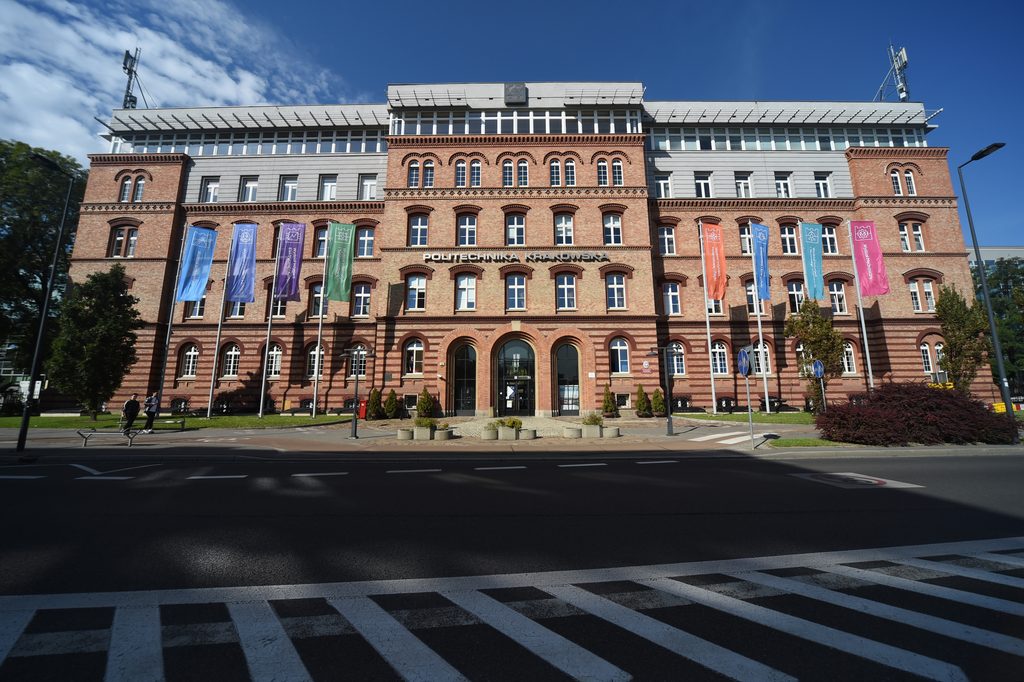 Budynek z brązowej cegły w stylu austro-węgierskim. wzdłuż fasady na wysokich masztach wiszą kolorowe flagi. To budynek główny Politechniki Krakowskiej.