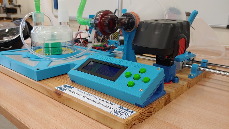 A colorful device—a portable respirator—placed on a wooden stand. In the corner of the stand is a white sticker featuring the navy blue logo of the Kraków University of Technology, the date, and the name of the artist: Damian Brewczyński