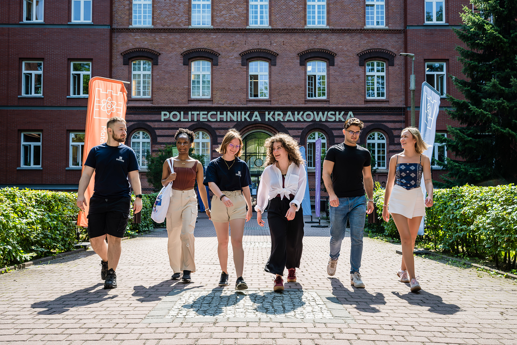 Six students, three girls and two boys. They are walking through the courtyard of Kraków University of Technology, laughing and talking. Behind them, the university's brick building and colorful faculty flags are visible.  