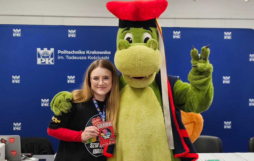 A person in a green dragon costume puts their arm around a young woman in a WOŚP T-shirt. Behind them, a blue wall with a white pattern of PK Krakow University of Technology signets is visible.