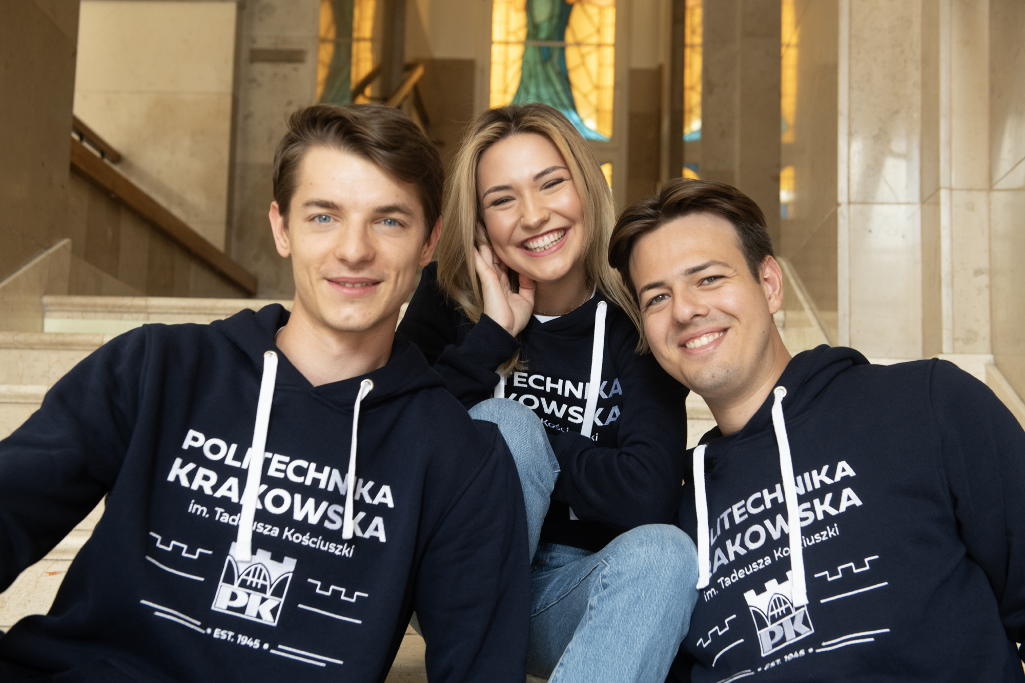 Dwóch studentów i jedna studentka. Siedzą na schodach, w tle widoczny jest witraż. Cała trójka ma na sobie granatowe bluzy z białym wyhaftowanym logo uczelni. Wszyscy się uśmiechają.