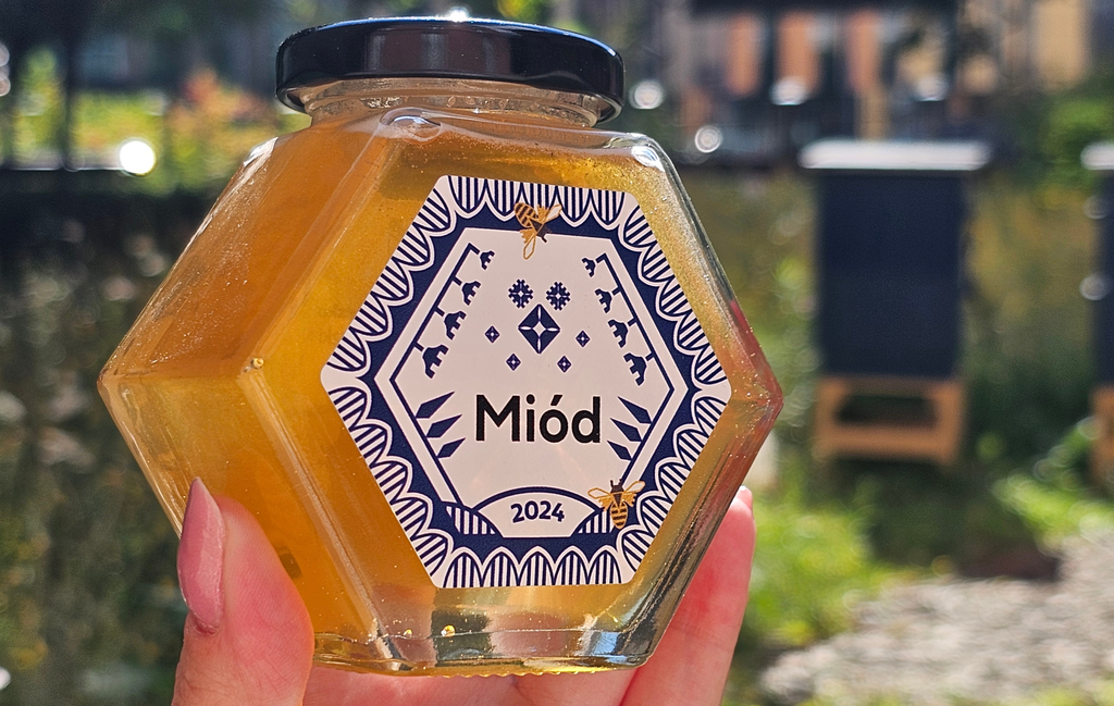 A jar of golden honey against the background of hives and a flowering meadow. A white and blue label on the jar.