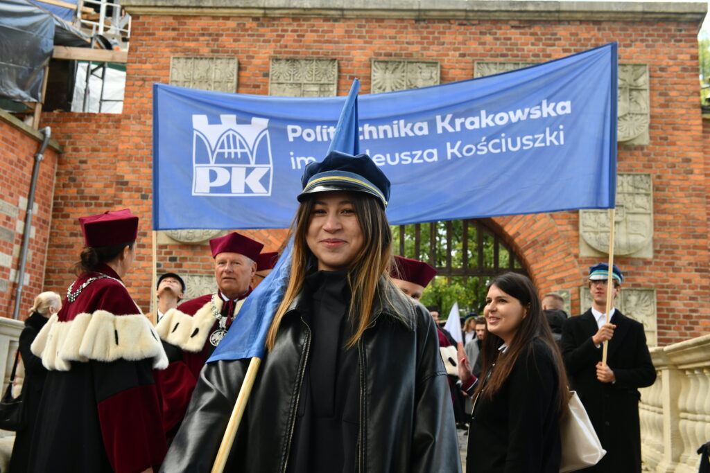 Młoda szatynka w ciemnym płaszczu i czapce studenckiej, trzyma niebieską flagę uczelni. Za nią pochód inauguracyjny i transparent: niebieski z białym logo Politechniki Krakowskiej