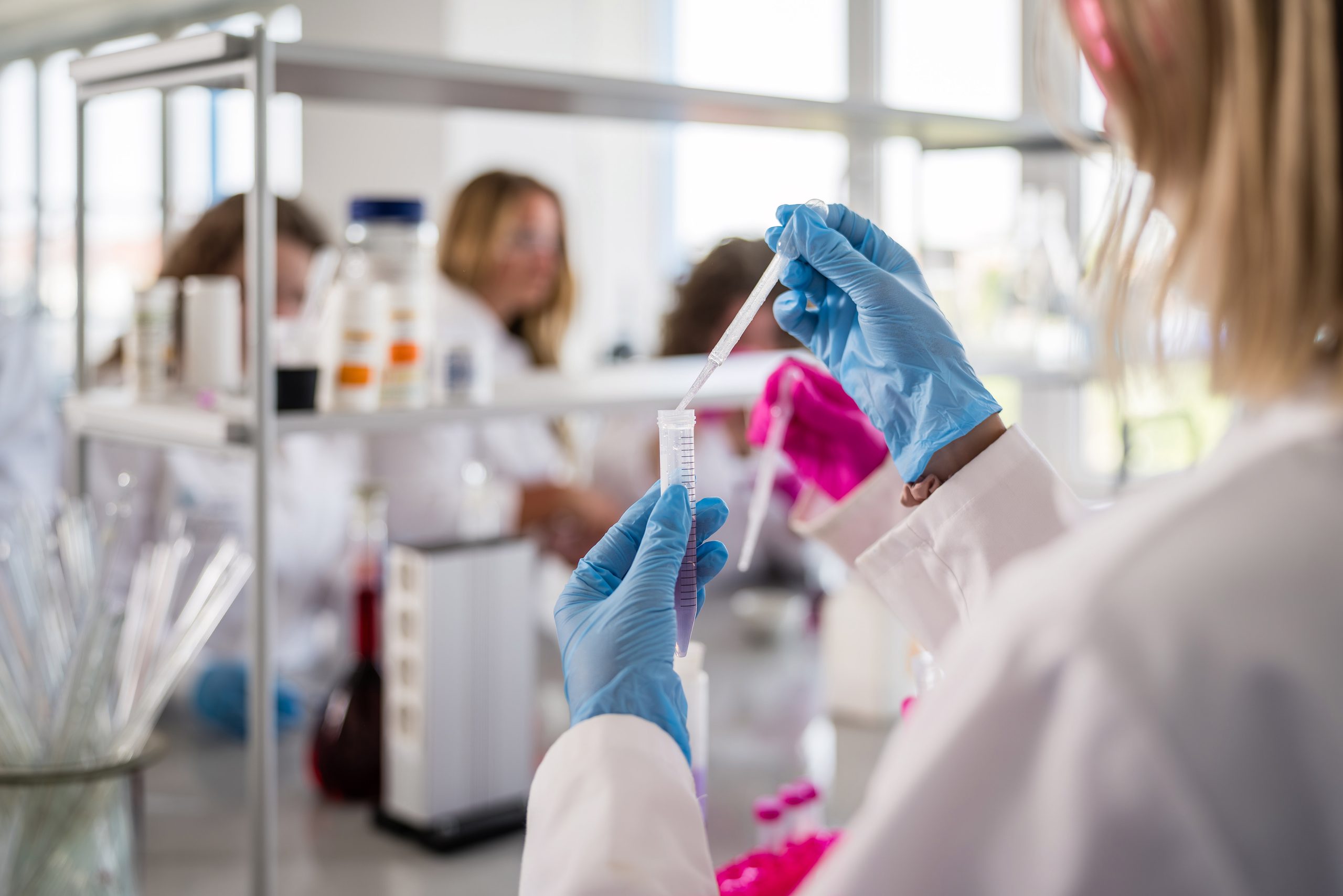 A laboratory. In the foreground, a woman with her back to the camera, wearing blue nitrile gloves. She is holding a flask and a pipette in her hands.