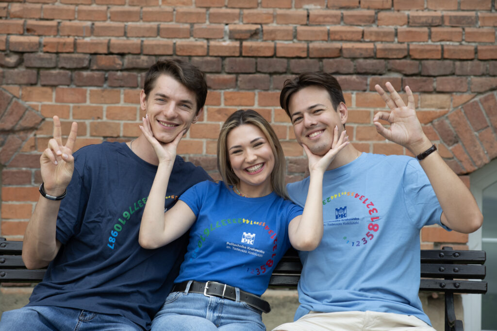 Three students: two boys and a girl between them. They’re smiling. They’re all wearing navy blue t shirts with the logo of the PK Kraków University of Technology embroidered on them