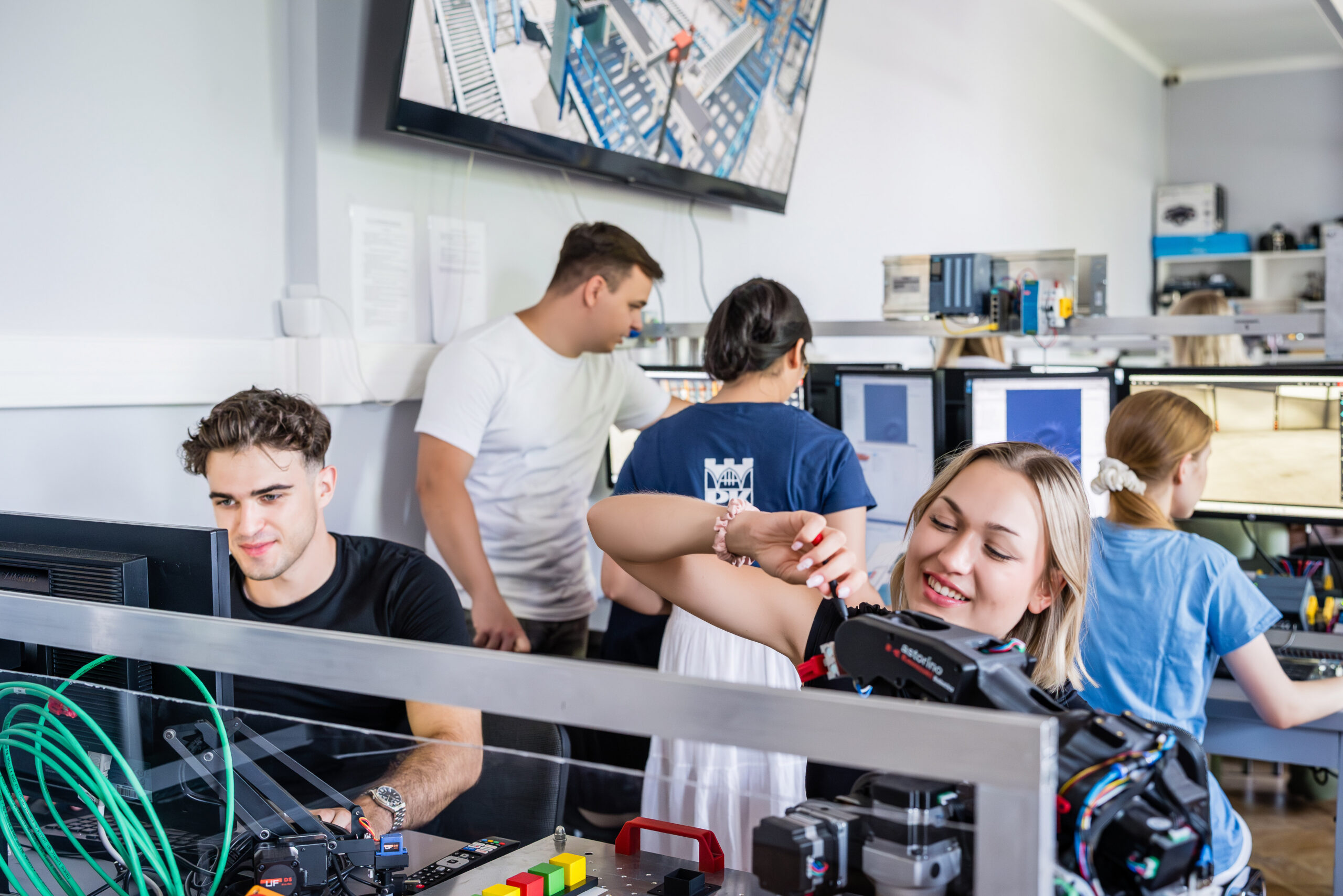 Grupa studentów przy stole do pracy z urządzeniami elektrycznymi. Przy pierwszym stanowisku: dziewczyna i chłopak. Ona trzyma śrubokręt i z uśmiechem skupia się na urządzeniu