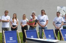 Grupa studentów w białych koszulkach stoi na tle szarej ściany przed nimi stoi młoda kobieta czarnej koszulce trzymająca w dłoniach bukiet kolorowych kwiatów. Spogląda w lewą stronę i się uśmiecha. Na dole stoją drewniane leżaki z granatowym materiałem i logo Politechniki Krakowskiej.